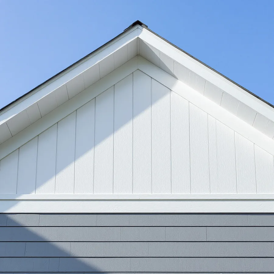 Looking up at a house gable clad in white vertical board and batten above gray horizontal lap siding, with a white trim band between the two