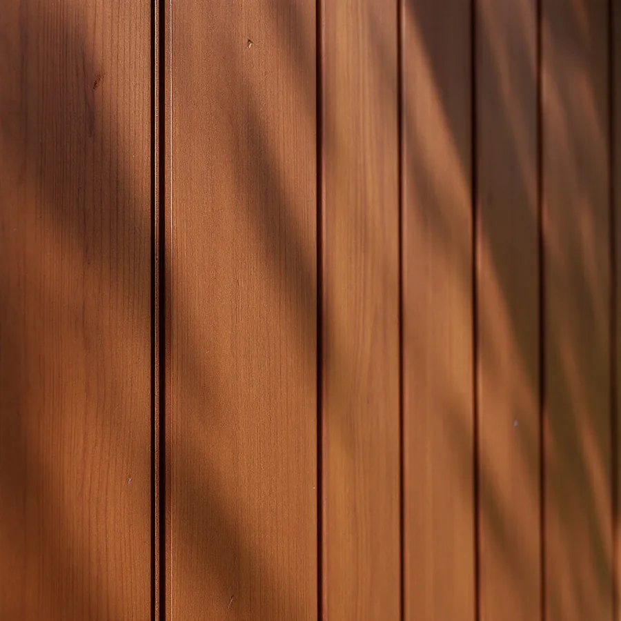 Close view of wide warm brown stained vertical boards separated by narrow dark recessed grooves, with dappled sunlight across the wall