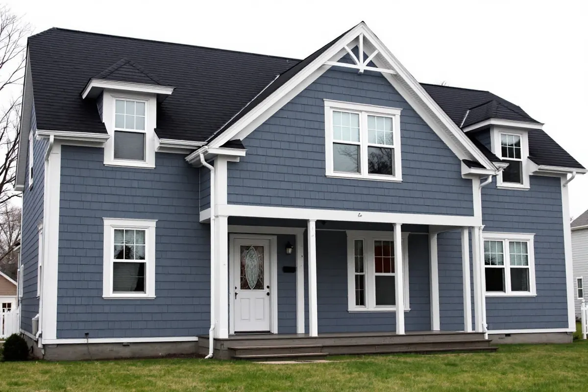 Two-story blue gray house with white trim, white porch posts and dormers, clad in painted siding under a dark shingle roof, with a lawn in front