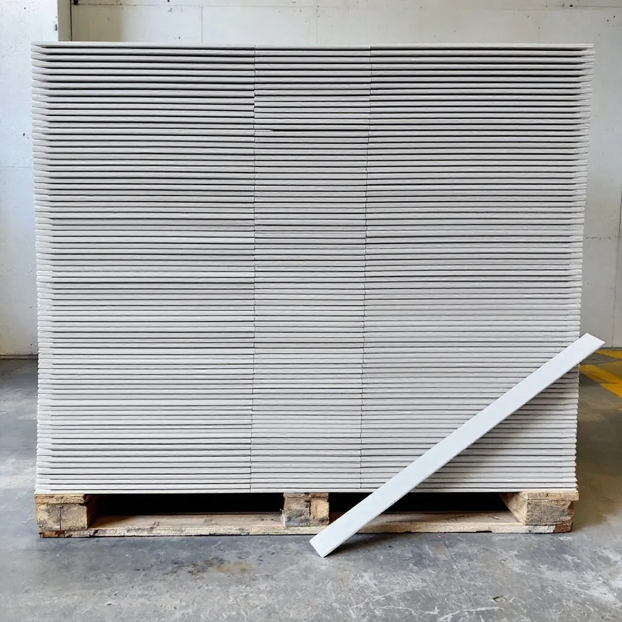 Tall stack of thin pale gray fiber cement planks on a wooden pallet on a concrete floor, one plank leaning against it