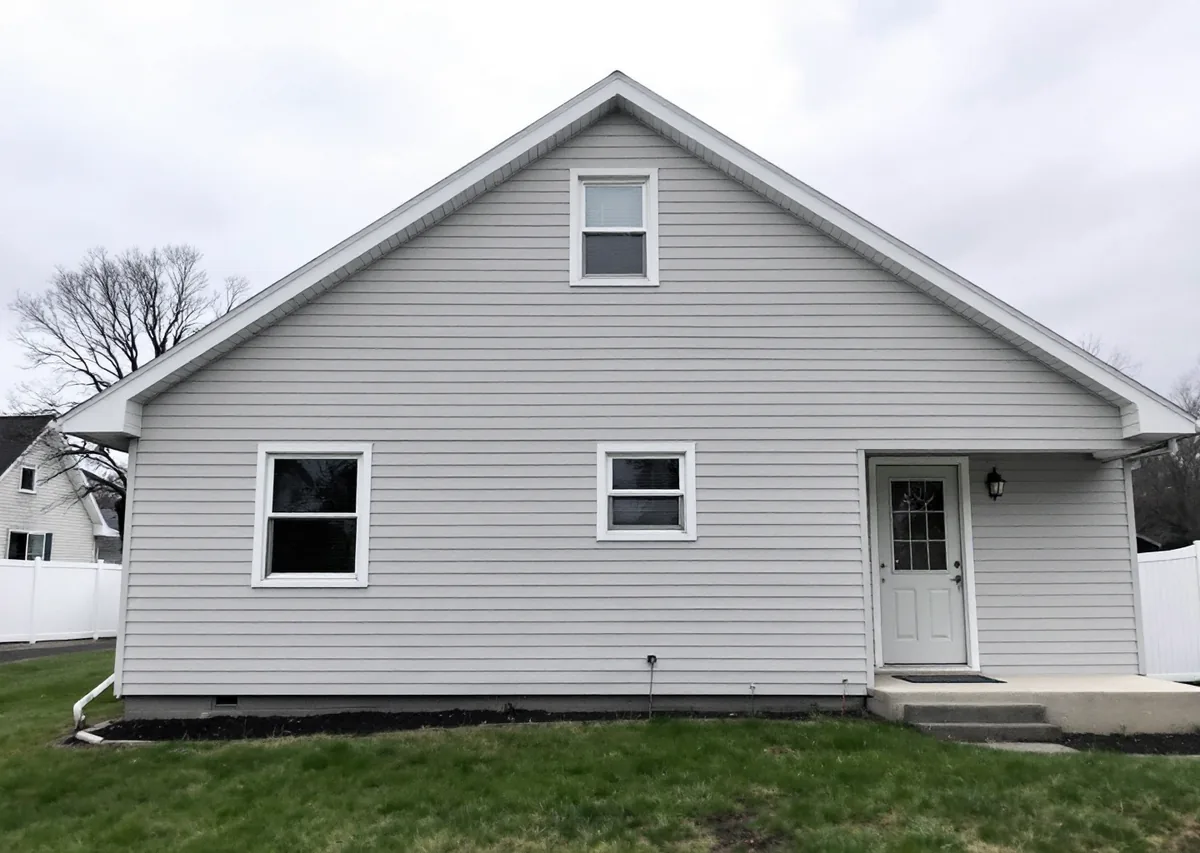 Straight-on view of the gable end of a two-story gray lap-sided house: a full triangular gable with one small window, two windows and a door with white trim on the ground floor, a lawn in front under an overcast sky