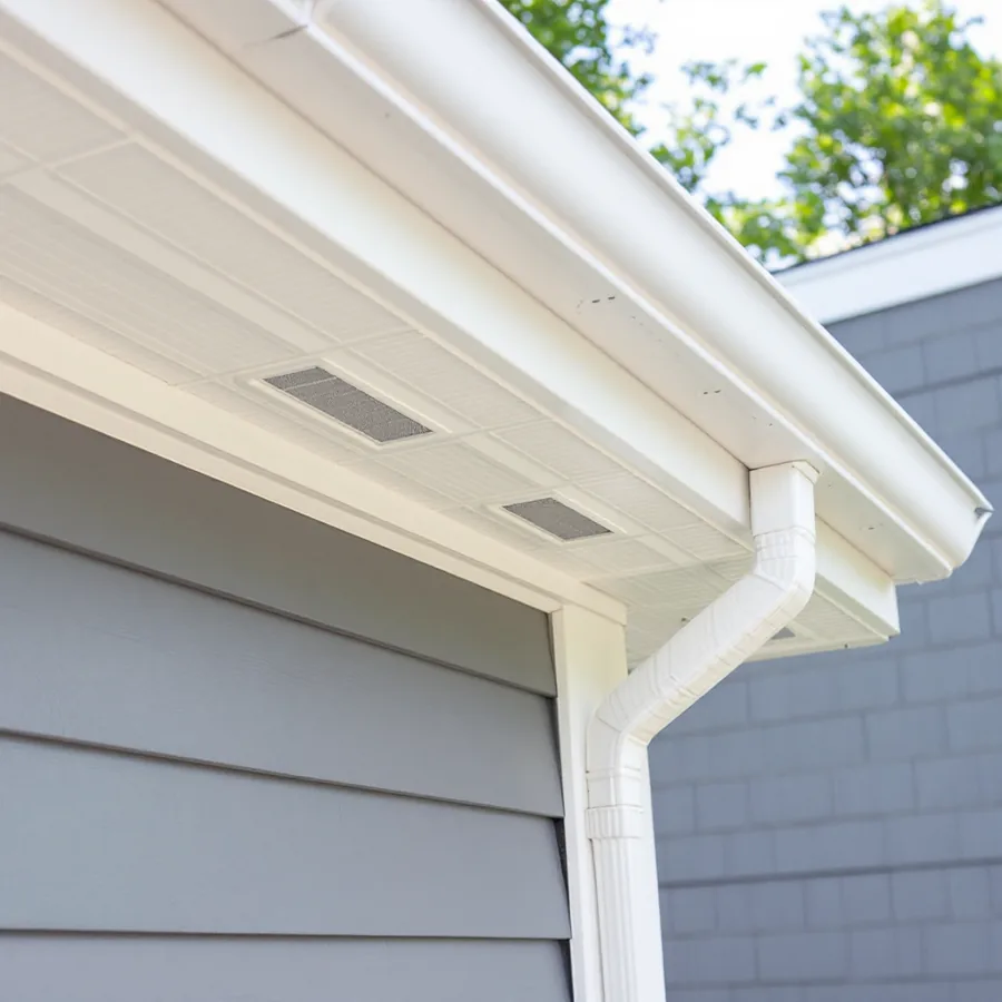 Looking up at a house eave with new white vented soffit panels, white fascia, gutter and downspout at the corner above gray lap siding