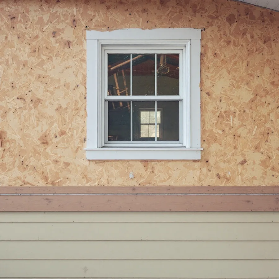 House wall with the old siding stripped off, showing bare OSB sheathing around a window and a strip of old siding at the bottom
