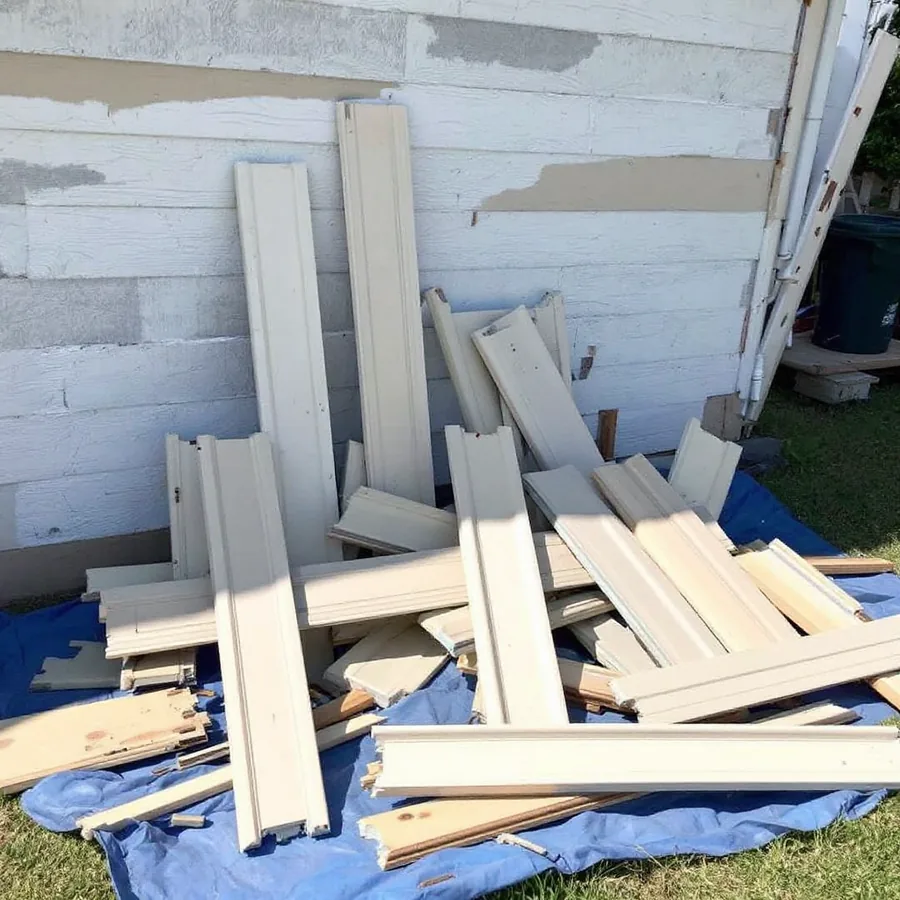 Pile of torn-off beige vinyl siding panels and broken trim on a blue tarp beside a stripped house wall